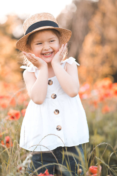 Cute funny baby girl 2-3 year old wear straw hat and white summer dress over flower meadow background. Kid laughing outdoors over nature. Looking at camera. Childhood.  - Photo, Image