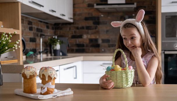 A beautiful child in a bandage with bunny ears, with colorful eggs in a basket, at home in the kitchen. Preparation for Easter - Fotó, kép