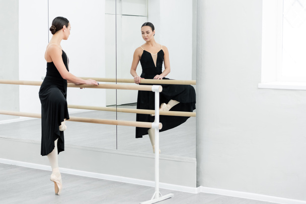 graceful ballerina in black dress training at barre near mirrors - Photo, Image