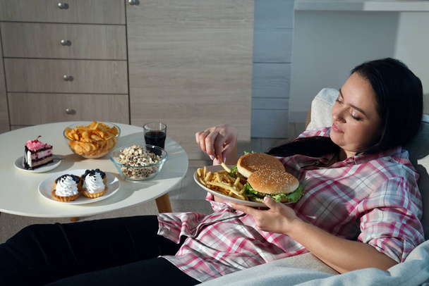 Happy overweight woman eating french fries with burgers at home - 写真・画像