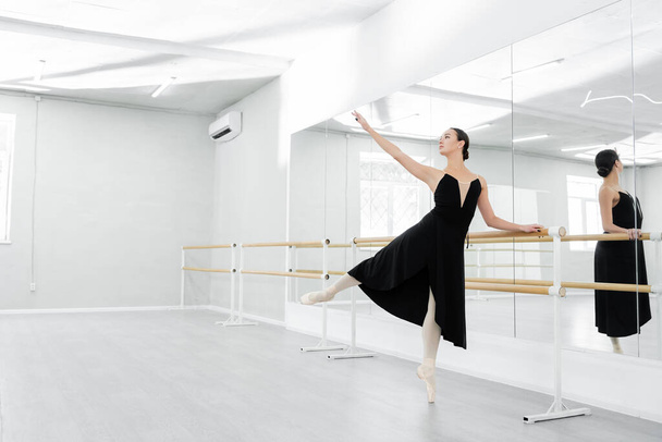 graceful woman in black dress rehearsing near mirrors in ballet studio - Photo, Image
