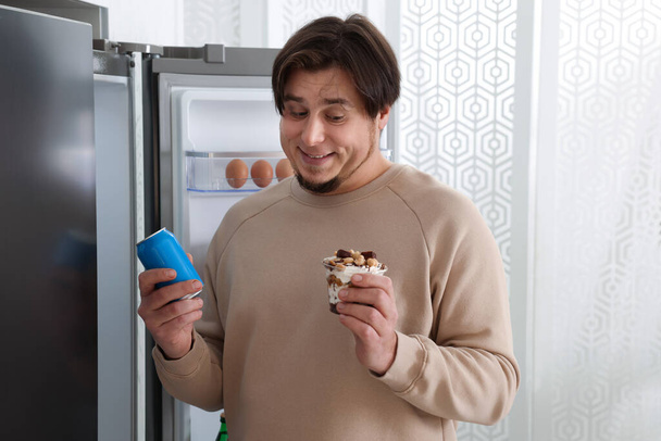 Overweight man holding dessert and tin can with beverage near open refrigerator in kitchen - Foto, immagini