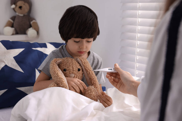 Doctor with thermometer near sick little boy in bed indoors - Foto, Imagen