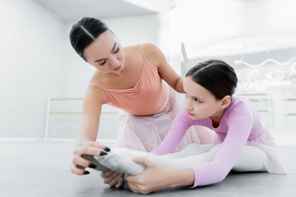 preteen girl looking at dance teacher while stretching on floor in ballet school - Fotó, kép
