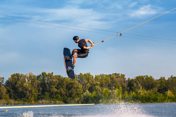 Skilled male wakeboarder jumping with backside rotation over water holding rope in hand - Photo, Image
