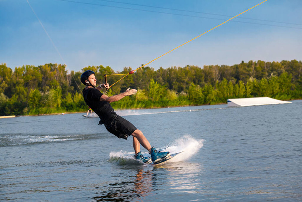 Hombre enfocado deslizándose en wakeboard en la superficie del agua sosteniendo la cuerda de remolque con una mano - Foto, Imagen