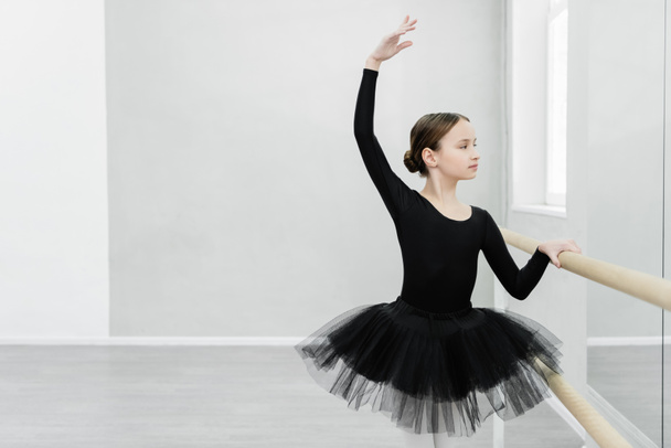 preteen girl in black tutu looking in mirror while training at barre in ballet studio - Foto, Imagen