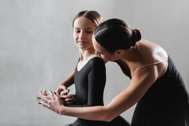 junge Ballettlehrerin berührt Hände von Mädchen während Tanzstunde auf grauem Hintergrund - Foto, Bild