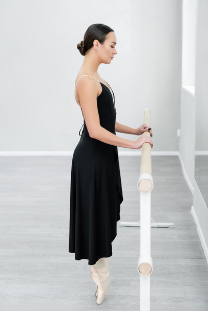boční pohled na baletní tanečnici v špičaté obuvi stojící na špičce u tyče ve studiu - Fotografie, Obrázek
