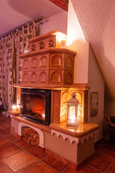 Beige tiled stove in the old style. Ceramic tiles in a cottage, vintage style of interior. Romantic night scenery with fireplace. View from angle. - Photo, Image