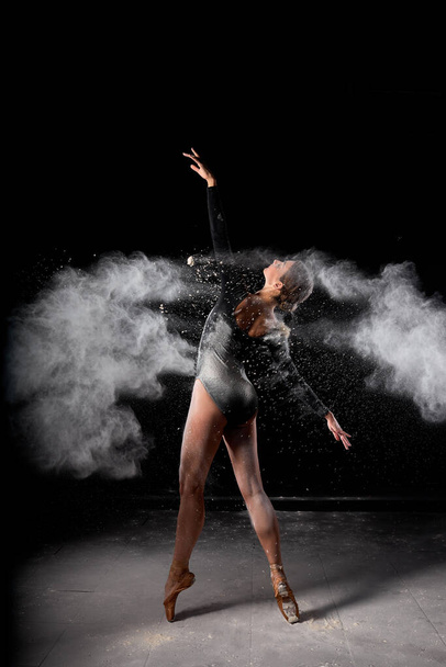 Beautiful pretty female wearing black gymnastic bodysuit dancing with flying powder - Foto, imagen