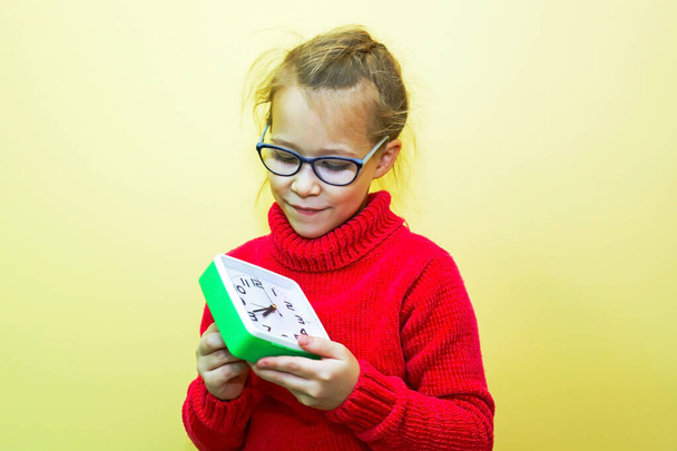 A child girl in a red sweater plays with an alarm clock, looks at the time and makes a gesture, showing the time or being late - Foto, Bild