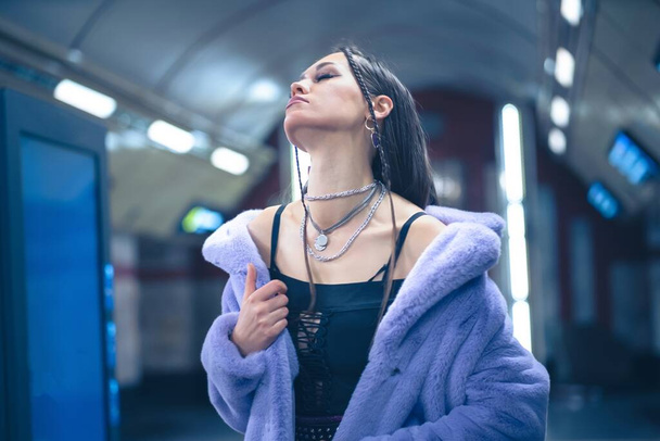 Sexy model in purple synthetic fur coat walking in metro or subway platform in blure background. Female with luxury makeup, long hair in stylish outfit - Foto, Imagem