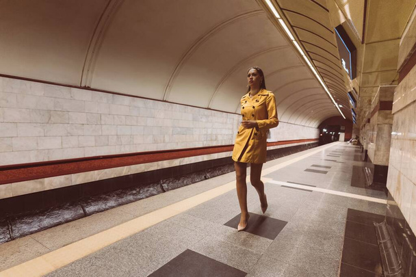Walking on the subway platform beautiful young girl with long legs in a yellow spring coat and a white handbag in her hand. - Photo, image