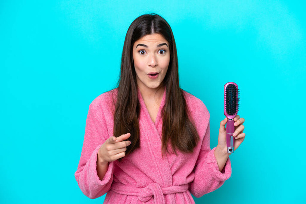 Mavi arka planda saç fırçası tutan genç Brezilyalı kadın şaşırmış ve ön tarafı işaret ediyor. - Fotoğraf, Görsel