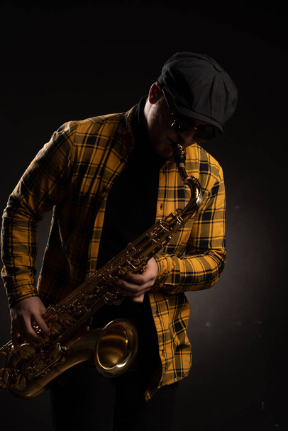 Stylish Tenor Sax Player in Yellow Plaid Shirt, Cap and Sunglasses Plays Saxophone on Black Background. Saxophonist Plays Jazz Music with Sax Alto. Close-up - Foto, Bild
