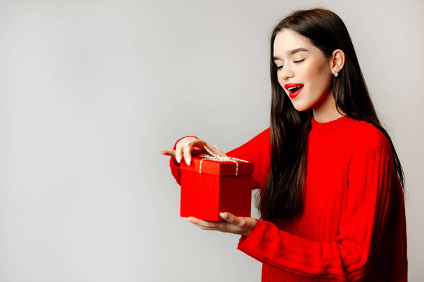 Beautiful young woman with black hair and red lips. The girl is staying against the light background and holding a present - Foto, Imagem