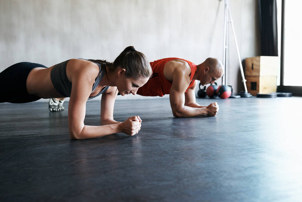 Empuje su camino a la aptitud. Tiro de un hombre y una mujer haciendo ejercicios de tablón en el gimnasio. - Foto, imagen