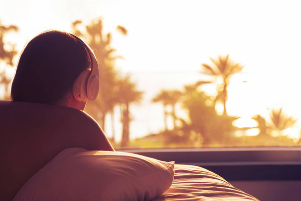 Silhouette of woman with headphones lying on pillow on the bed in bedroom opposite panoramic window with view on palm tree sea beach at sunset. Female relaxing listenning music. - Foto, Imagem