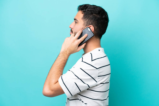 Joven brasileño aislado sobre fondo azul manteniendo una conversación con el teléfono móvil con alguien - Foto, Imagen