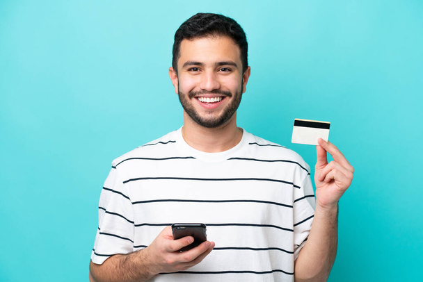 Junger Brasilianer isoliert auf blauem Hintergrund kauft mit dem Handy mit Kreditkarte - Foto, Bild