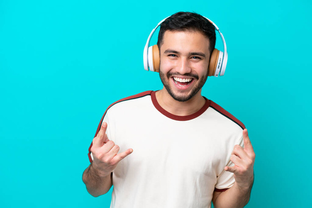 Fiatal brazil férfi elszigetelt kék háttér hallgat zenét, hogy rock gesztus - Fotó, kép