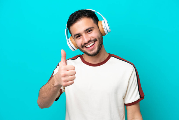 Nuori brasilialainen mies eristetty sinisellä taustalla kuunteleva musiikki ja peukalo ylöspäin - Valokuva, kuva