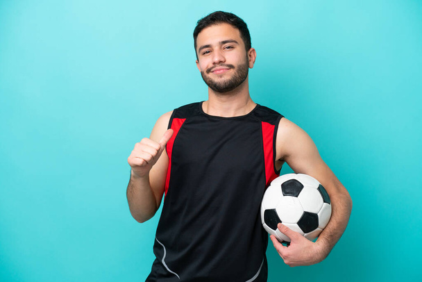 Young football player Brazilian man isolated on blue background proud and self-satisfied - 写真・画像