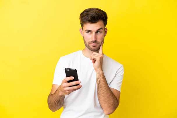 Genç, beyaz, yakışıklı bir adam sarı arka planda cep telefonu kullanıyor ve düşünüyor. - Fotoğraf, Görsel