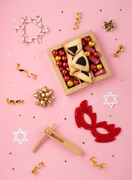 Purim bayramı Yahudi karnavalı konsepti. Lezzetli hamantaschen kurabiyeleri, kırmızı karnaval maskesi, gürültü çıkarıcı, tatlı şekerler ve pembe arka planda parti dekoru. Üst görünüm, düz görünüm, kopyalama alanı. - Fotoğraf, Görsel