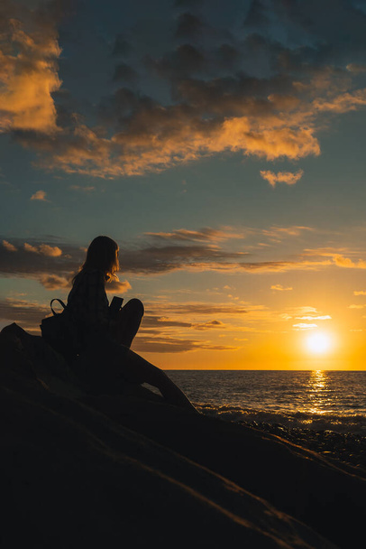 若い女性のシルエットは海岸に巨大な石の上に座って、海に行く太陽と美しく、魔法のような、明るい夕日を見ています。垂直写真 - 写真・画像