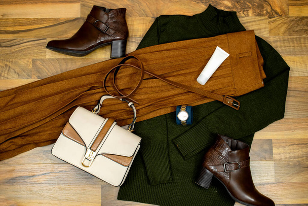 ファッショナブルな服やアクセサリーのセット。緑のセーターと段ボールスカート、ハンドバッグ、足首のブーツと茶色の革ベルトと木製のテーブル、フラットレイアウト上のチューブ顔クリーム。傾向にある - 写真・画像
