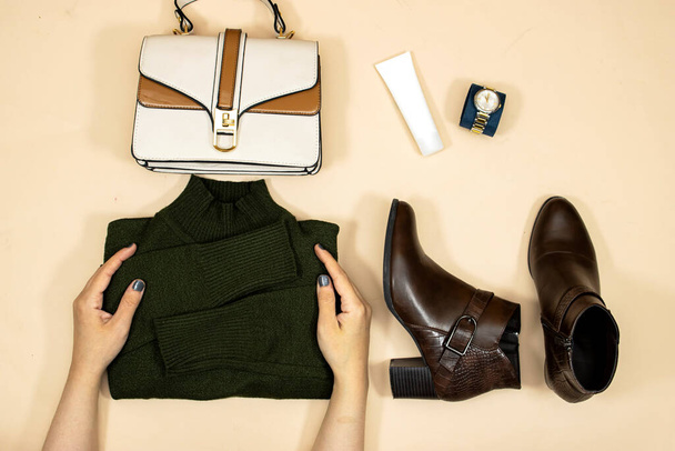Un conjunto de ropa y accesorios de moda. manos mujer sosteniendo suéter verde y tubo crema cara, bolso, botas de tobillo y reloj de mano, Flat lay. Estar en tendencia - Foto, Imagen