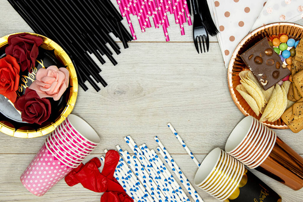 concept joyeux anniversaire, chocolat bonbons et paille et croustilles pomme de terre, biscuits et tasses jetables avec fourchettes en plastique et roses dans une assiette en papier sur table en bois - Photo, image