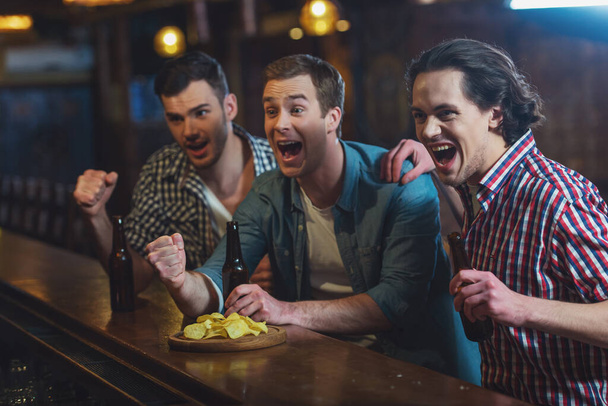 Three young men in casual clothes are cheering for football and holding bottles of beer while sitting at bar counter in pub - Фото, изображение
