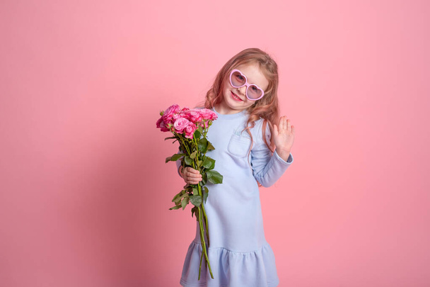 cute child girl in blue dress and sunglasses holding bouquet of pink roses on pink background - Fotografie, Obrázek
