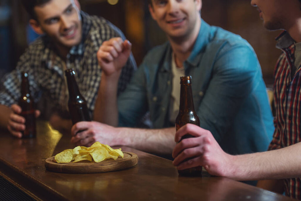 Kolme nuorta miestä tavallisissa vaatteissa puhuvat, syövät sipsejä ja juovat olutta istuessaan baaritiskillä pubissa, lähikuva
 - Valokuva, kuva