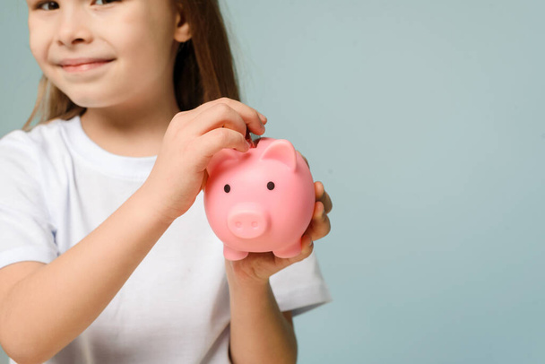 A little girl puts a coin in a piggy bank. The concept of teaching children personal finance and saving. - Photo, Image