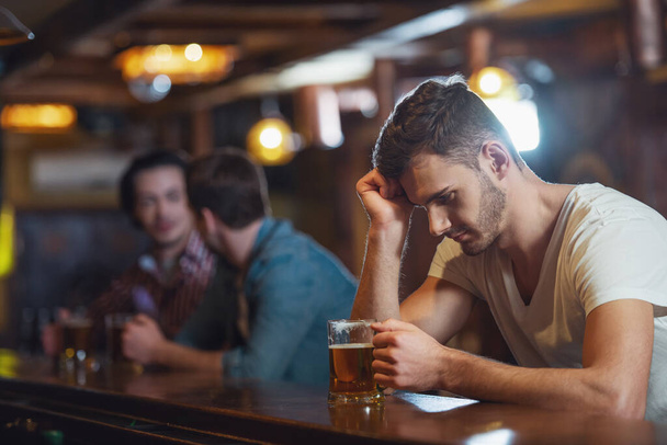 Jovem triste em t-shirt branca está olhando para o copo de cerveja enquanto sentado no balcão do bar no pub, no fundo dois outros homens
 - Foto, Imagem