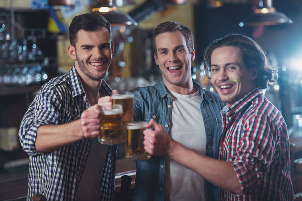 Three young men in casual clothes are smiling, looking at camera and clanging glasses of beer together while standing near bar counter in pub - Photo, Image