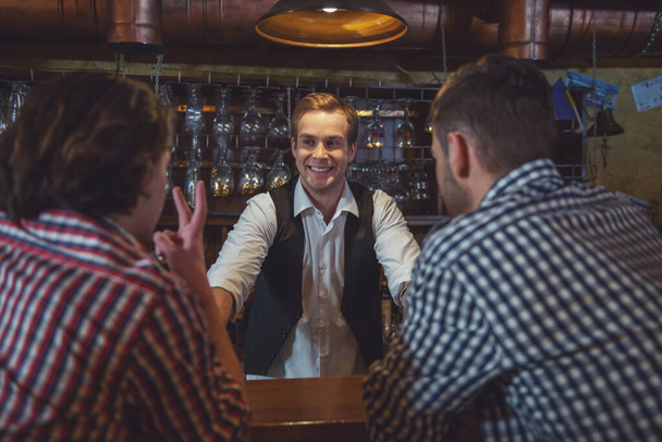 Barman atraente está sorrindo e tomando a ordem de dois homens sentados no balcão do bar no pub
 - Foto, Imagem