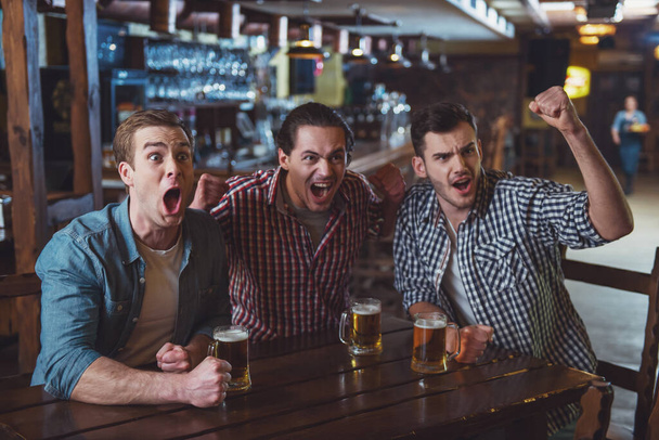 Tres jóvenes con ropa casual están animando al fútbol y sosteniendo vasos de cerveza mientras están sentados en el pub.
 - Foto, Imagen