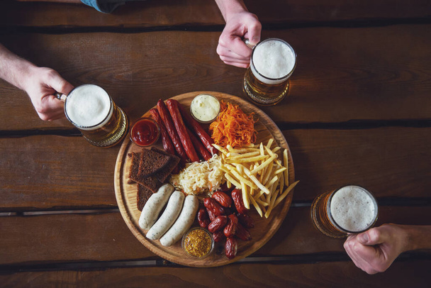 Top view of hands holding glasses of beer, a big wooden tray with delicious snacks near, on wooden background - Photo, Image