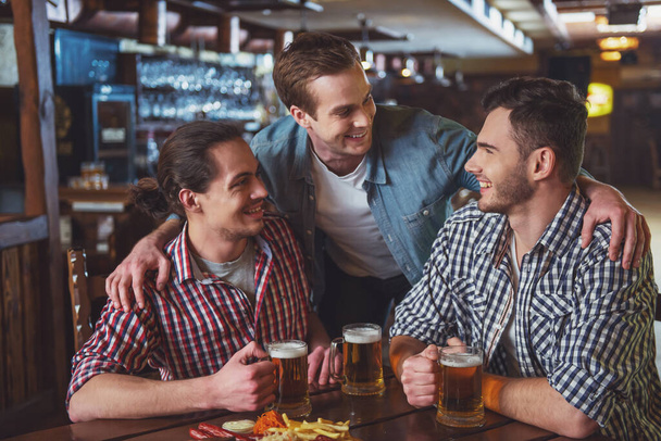 Three young men in casual clothes are smiling, talking and drinking beer while sitting in pub - Fotoğraf, Görsel