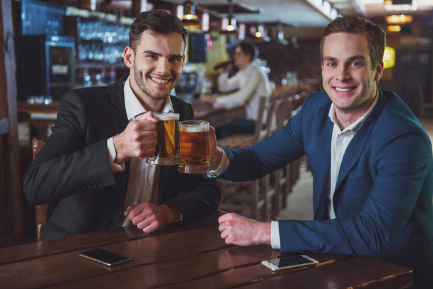 Dos jóvenes hombres de negocios con trajes sonríen, mirando a la cámara y colgando vasos de cerveza mientras están sentados en el pub
 - Foto, imagen
