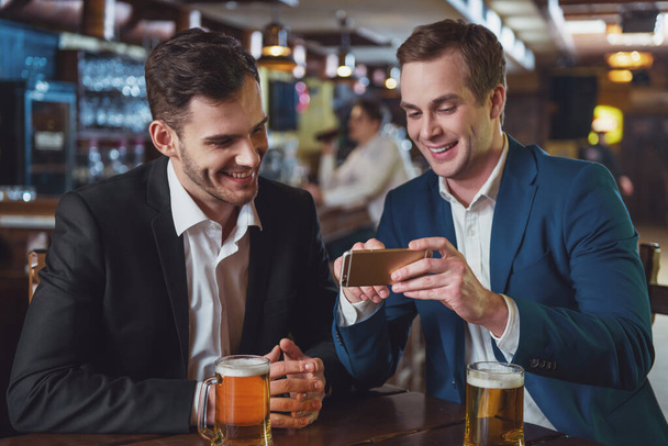 Dos jóvenes hombres de negocios con trajes sonríen, usando un teléfono móvil y bebiendo cerveza mientras están sentados en el pub
 - Foto, imagen