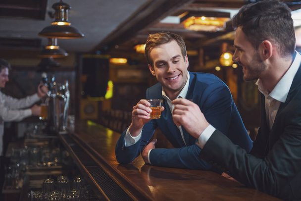 Two young businessmen in suits are smiling and clanging glasses of alcoholic beverage together while sitting at bar counter in pub - Foto, Imagem