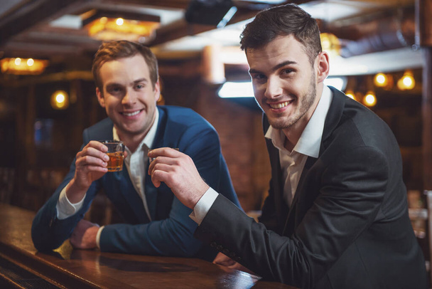 Two young businessmen are smiling, looking at camera and clanging glasses of alcoholic beverage together while sitting at bar counter in pub - Foto, Imagem