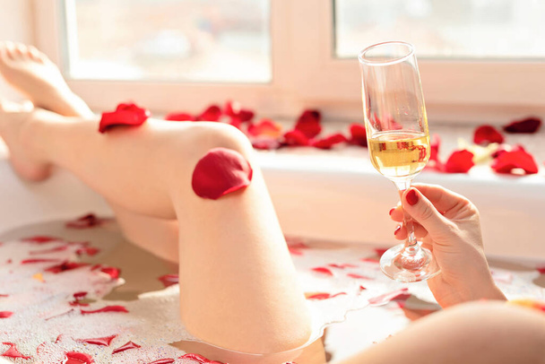 Female hand holding a glass of white wine while sitting in a bathtub with foam and red rose petals. Enjoying life concept. Selective focus - 写真・画像