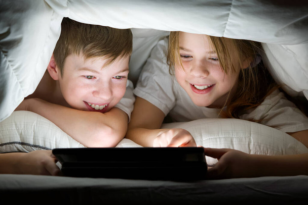 kinderen spelen 's nachts spelletjes op een tablet onder een deken, kinderen kijken video op tablet. communicatie via sociale netwerken. - Foto, afbeelding